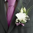 A close-up wedding portrait of a man's charcoal grey suit jacket shows a single white lisianthus buttonhole glowing softly against the dark fabric, the detail so sharp you can almost feel the textures. The fine wool suit, with its subtle woven pattern and crisply pressed lapel, is paired with a deep magenta, almost fuchsia, silk tie that runs like a ribbon of colour down the centre of the frame. Beneath the tie, a pale grey and white patterned dress shirt peeks through, its tiny geometric print adding a quiet sophistication. Pinned neatly to the left lapel is the boutonnière: one fully open white lisianthus bloom, ruffled petals layered like delicate tissue, surrounded by two slender ivory buds just beginning to unfurl. Airy clouds of white gypsophila dance around the main flower, while fine, linear greenery arches outwards, giving the arrangement a fresh, just-picked feel, as if gathered from a garden near Stanmore Country Park on a cool spring morning. At the base, a sleek green leaf wraps the stems and is finished with a narrow mauve ribbon, tied carefully to echo the rich tones of the tie without overwhelming the design. The lighting is soft and even, with no harsh shadows, so every petal edge, stitch in the lapel, and fold in the silk knot appears clear and intentional. The composition conveys the quiet excitement of a groom dressing for a ceremony in Stanmore, perhaps at a local church off Marsh Lane or a reception near Bentley Priory, and reflects the thoughtful craftsmanship a florist brings when creating an understated, romantic buttonhole for a day filled with hope, promise, and new beginnings.