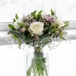 An elegant white rose bouquet stands in a simple cylindrical glass vase, bathed in soft daylight that feels like it could be streaming through a quiet Stanmore bay window. Three fully open, creamy white roses form the calm heart of the design, each bloom with satin-smooth petals gently curling outward to reveal a faint ivory centre, suggesting absolute freshness. Around them, rich emerald greenery is layered with care: broad, slightly folded leaves cradle the roses, while smaller textured foliage adds depth and a natural, garden-like fullness. Threaded between the roses are sprays of delicate white gypsophila, or baby's breath, their tiny star-shaped flowers floating like mist and giving the bouquet a light, airy halo. Subtle touches of colour arrive through alstroemeria buds and blossoms in muted mauve and blush-pink tones, their slender petals streaked and speckled, hinting at romance without overpowering the purity of the white roses. The stems sit clearly visible through the transparent vase, neatly gathered and tied just above the waterline with a fine, unobtrusive ribbon, showing a florist's practiced hand. Clear water fills the vase, catching the light and amplifying the fresh green of the stems. The background is softly blurred and luminous, a pale white that suggests a bright morning in a Stanmore home looking out towards Bentley Priory Nature Reserve. The overall composition is rounded, compact, and harmonious, evoking a sense of serenity and understated luxury. It feels perfectly suited to a quiet wedding, a condolence message, or a thoughtful gesture of support, the kind of classic white and green arrangement a local Stanmore florist might recommend for moments that call for gentle beauty and calm reflection.