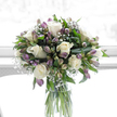 Standing in a simple clear glass cylinder vase, this sympathy bouquet carries the quiet eloquence of a floral tribute prepared by Flowers Stanmore for a family gathering in a peaceful home near Canons Park. At the centre, several creamy white roses open fully, their petals smooth and softly layered, forming luxurious cups that communicate purity, love, and gentle remembrance. Weaving between and around these roses, alstroemeria blooms in subtle shades of lilac, lavender, and deepened purple, with faint hints of green at the petal bases, recalling the restrained colours of a shaded Stanmore garden. Some of these blooms remain as closed buds, promising gradual unfolding, while others are half-open, adding texture and a sense of quiet progression. Fine sprays of white baby's breath are scattered throughout, forming a delicate lace of tiny florets that fill any empty spaces and impart an airy, almost weightless quality to the arrangement. The bouquet is framed by lush green foliage, composed of glossy leaves and more slender, textural greens that curve gently around the flowers, offering both support and visual depth. The stems are clearly visible through the vase, standing straight in fresh water, reinforcing a feeling of care and order. Behind the arrangement, a bright, softly blurred white background suggests diffused daylight streaming through a window on Stanmore Hill, highlighting the bouquet's clean lines and the softness of its colours. This gentle interplay of whites, purples, and greens makes the design particularly suited for expressing sympathy or quiet remembrance, bringing a calm, dignified presence to homes across Stanmore, from leafy residential roads off The Broadway to more secluded properties near Stanmore Country Park.