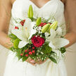 In this image, a hand-tied bridal bouquet is held close to a lace wedding gown, the flowers and dress forming a beautifully layered tableau that feels perfectly suited to a Stanmore wedding. Generous white lilies stretch out their creamy, star-shaped petals, some fully open, others just beginning to unfurl, each one casting soft shadows that emphasise their curves. At the centre, a single red rose stands out like a glowing ember, its deep crimson petals densely folded and luxuriously velvety, anchoring the design with a classic symbol of passionate love. Fine sprays of baby's breath drift between the larger blooms, their tiny white flowers echoing the delicate patterns of the lace beneath. Dotted among the petals, glossy red berries and clusters of hypericum add lively touches of colour, lending the arrangement a festive, joyful energy that might suit a reception near Stanmore Hill or close to the station. Mossy green textures at the base and tufts of bright green dianthus contribute both softness and structure, suggesting the freshness of nearby open spaces and gardens. Slender strands of ornamental grass arc outwards in airy sweeps, giving the bouquet a graceful, almost floating quality, as though it might sway gently with each step the bride takes down the aisle. The stems are bound in ribbon, neatly finished and partly revealed beneath the bride's fingers, which rest around the bouquet with gentle confidence. Natural light bathes the scene, highlighting the contrast between the smooth lily petals, the plush rose, and the intricate lace. The resulting impression is one of refined romance and careful craftsmanship, a bouquet that marries tradition with a light, contemporary touch for weddings across the Stanmore area.