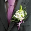 A close-up, high-angle photograph captures the upper chest of a man dressed for a formal event in Stanmore, his attire centred around a charcoal grey suit jacket with clean, structured tailoring. The lapel lines are sharp and confident, framing a pale dress shirt patterned with a subtle grid that adds quiet texture rather than distraction. Down the centre runs a smooth lavender tie, neatly knotted and lying flat, its soft sheen catching a hint of daylight as you might see outside a ceremony at a Stanmore synagogue or a registry office near the Broadway. Pinned precisely to the left lapel is a refined wedding buttonhole: a tall spire of pale green buds, resembling an unopened Muscari or hyacinth, rises elegantly at the top, suggesting freshness and anticipation. Nestled around this focal stem, several white alstroemeria blooms open fully, each petal marked with fine maroon streaks and a whisper of gold at the stamens, like tiny brushstrokes against porcelain. Sprays of white gypsophila weave through the design, their cloud of tiny flowers softening the edges and creating a romantic halo. Slender, grass-like blades loop gracefully alongside broader, deeper green leaves, giving movement and contrast against the dark wool of the suit. At the base, a lavender satin ribbon, perfectly colour-matched to the tie, is tied into a small bow that gathers the stems together with understated precision. The whole composition feels calm, polished, and quietly joyful, the kind of carefully coordinated boutonniere a Stanmore groom or prom guest might choose to mark an important milestone with elegance and a touch of local charm.