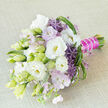 A sophisticated hand-tied bouquet is captured resting at a gentle angle on a softly textured, light beige fabric, the neutral backdrop highlighting every tonal nuance of white, lavender, and green. The composition is dominated by velvety white lisianthus blooms, some fully unfurled to reveal creamy yellow throats and delicately ruffled petals, others still closed as tapered buds in shades of pale green and ivory, hinting at blossoms yet to open. Around them, small clusters of pastel lavender-pink flowers create a cloud-like, airy layer, softening the structure and adding a romantic haze, much like the soft evening light over Stanmore's leafy side streets. Striking purple allium florets, each made of tiny star-shaped flowers forming a textured sphere, provide a dramatic counterpoint to the lisianthus, their spiky silhouettes adding a touch of contemporary flair. Varied foliage weaves through the bouquet: slender, elongated leaves, softly looping strands of grass, and lighter green, spiky accents that contribute both movement and subtle contrast. The stems at the base are neatly bound to form a comfortable handle, wrapped in a bold fuchsia satin ribbon that gleams against the neutral background. A fine strand of white pearls is coiled over the ribbon, giving the handle an elegant, jewellery-like finish reminiscent of bridal accessories seen at weddings in and around Stanmore. The overall mood is one of purity, refined romance, and quiet celebration, as though this bouquet is ready to be carried into a ceremony at a local venue or delivered as a bespoke gift from a trusted Stanmore florist, bringing with it a gentle fragrance and a sense of thoughtful craftsmanship.