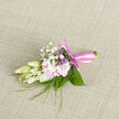 Resting on a softly woven, neutral fabric that could easily be found in a cosy Stanmore townhouse, this small boutonnière reveals a careful symphony of texture and tone. At the centre, creamy buds, likely delicate freesias, sit just on the verge of opening, their petals almost translucent at the edges as they catch the gentle light. They are closely paired with pale pink blooms that unfurl with a soft, sighing elegance, providing a tender blush of colour. Tiny tufts of white gypsophila, like floating clusters of miniature clouds, weave in between the main flowers and add an intricate lacework effect that lightens the composition. Slender, blade-like greenery arcs gracefully outwards, complemented by broader glossy leaves that cradle the arrangement, giving it a sense of structure and poise. Everything is bound together by a smooth, satin pink ribbon, wrapped tightly around the stems and finished in a neat, precise knot that shows the florist's practiced hand. You can imagine a gentle perfume-fresh, sweet, and unobtrusive-lingering softly in the air, reminiscent of gardens around Stanmore on a mild spring afternoon. This boutonnière feels perfectly suited for a wedding or prom, perhaps worn at a celebration near Stanmore station or along The Broadway, its subtle colour palette adding a quiet touch of refinement to a suit. Prepared with the same care that Flowers Stanmore brings to all local arrangements, it radiates intimacy, composure, and a sense of lovingly crafted detail meant for cherished moments.