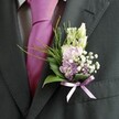 A detailed close-up photograph focuses on the upper body of a man dressed for a formal occasion in Stanmore, showing only his charcoal grey, almost black, suit jacket, pale patterned shirt, and bold magenta silk tie. The fabric of the jacket has a subtle texture that catches the soft daylight, especially along the lapel where a meticulously crafted wedding boutonnière is pinned. The tie, a rich fuchsia-pink with a smooth, lustrous finish, stands proudly at the centre, its knot precise and balanced, echoing the confident mood of a groom or honoured guest arriving at a ceremony near Stanmore Country Park. The floral detail on the lapel forms the true focal point: at the top, slender, unopened buds of creamy white or pale ivory flowers-resembling snapdragons-rise gently, suggesting blooms just on the edge of opening. Nestled beneath them is a cluster of soft pink and delicate lavender-purple blossoms, reminiscent of sweet peas or stock, mixed with a few slightly deeper purple florets that add quiet depth. Scattered between these petals are tiny stars of white gypsophila, or baby's breath, which create a light, misty veil around the central flowers. At the base, glossy dark green leaves provide structure, while fine, feathery strands of brighter greenery arc outward like small fern fronds, giving the arrangement movement and a garden-fresh feel, as if it had just been gathered from a hidden corner of Bentley Priory Nature Reserve. A neat lavender satin ribbon is tied in a small bow around the stems, its soft sheen echoing the lilac tones in the flowers and securing everything in place. Together, the bold tie and the nuanced pastel buttonhole convey an elegant yet playful sense of celebration, perfectly suited to a Stanmore wedding morning.