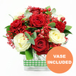 This photograph features an elegant rose arrangement that feels perfectly at home in Stanmore, perhaps adorning a dining table after a leisurely walk through Canons Park. The bouquet is composed of velvety deep red roses and creamy white roses, each bloom fully formed, with petals gently curling outward to create soft, layered cups of colour. The rich red and calm white tones play against one another, giving the display a romantic, slightly festive character. Threaded between the main flowers are delicate tufts of bright red filler blossoms, reminiscent of finely clustered gypsophila, which create a misty, airy texture, as if the bouquet were gently dusted with tiny red stars. Glossy red hypericum berries are scattered throughout, their round, bead-like shapes gleaming among the petals and adding both depth and a sense of abundance. Encircling the roses, lush greenery spreads out in varied shades of green, with slender eucalyptus sprigs offering a cool, silvery accent and other foliage providing structure and fullness, like a frame of fresh garden leaves cut on a crisp Stanmore morning. The stems are gathered in a clear, rectangular glass vase, its transparent sides revealing the straight lines of the stems and the water within, contributing to a modern, orderly look. Around the middle of the vase, a neatly wrapped green and white gingham ribbon adds a cheerful, rustic touch that suggests home-cooked meals and family gatherings. In the lower right corner of the image, a vibrant orange circular icon reads VASE INCLUDED in clear white text, making it obvious that this is a complete, ready-to-display floral gift, well suited to anniversaries, Valentine's Day, or simply sending a heartfelt message to someone in Stanmore with fresh, beautifully arranged roses.