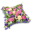 The photograph features a sumptuous, cushion-shaped floral tribute, its soft rectangular outline evoking the idea of a comforting pillow placed with care to honour a loved one. Positioned against a clean white background, the arrangement stands out in vivid detail, as if ready to be delivered to a quiet family home or chapel in Stanmore by the team at Flowers Stanmore. The tribute is entirely covered with fresh blooms, dominated by generous roses in a palette of fuchsia, blush pink, and creamy pale yellow. Some roses are fully opened, their swirling, layered petals creating plush, velvety rosettes; others are more tightly furled, adding depth and a sense of gentle movement across the cushion's surface. Interspersed among these are graceful deep purple lisianthus flowers, their ruffled petals bringing a luxuriant, velvety note that deepens the overall colour scheme and offers a dignified contrast. Smaller blush pink spray roses nestle into spaces between the larger flowers, creating pockets of delicate detail and warmth. Throughout the design, clusters of tiny lavender-hued filler flowers, likely statice or limonium, provide fine, airy texture and a misty touch of colour, a little like the distant haze over the trees around Canons Park. Encircling and underpinning the entire tribute is an abundance of foliage: rounded, silvery-green eucalyptus leaves lend a cool, soothing tone and a softly powdery texture, while darker, fine, fern-like greenery creates shadows and structure, ensuring the floral pillow looks full and dimensional. The overall feeling is peaceful, tender, and respectful, making this cushion arrangement a meaningful choice for funerals, memorial services, or graveside tributes in Stanmore, where families wish to express sympathy, gratitude, and enduring love through beautiful, carefully crafted flowers.