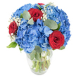 A bright, tactile bouquet of cobalt-blue hydrangeas and three velvety red roses sits in a simple clear glass vase, its rounded base and gently tapered neck visible against a clean white backdrop. The hydrangeas form a plush, cloud-like mass of tiny overlapping petals, their sky-blue faces dense and saturated as if the summer sky were gathered into stems. Nestled into this blue cloud are three deeply red roses, their petals still tightly spiralled and soft to the touch, providing a striking, intimate counterpoint that reads as both romantic and celebratory. Fine sprays of white baby's breath (Gypsophila) float between the larger blooms like pale confetti, catching the light and giving the arrangement an airy, ephemeral texture. Around and beneath the blossoms, muted sage-green eucalyptus leaves - rounded, slightly silvered, and smooth - weave through the composition, adding depth, a subtle evergreen scent, and a calming visual coolness. The glass vase reveals clean green stems submerged in clear water, a small honest detail that speaks of recent conditioning and freshness. Bright, natural lighting lifts the colours - the blue appears almost electric, the red rich and velvety - and casts soft, delicate shadows across the white surface. As a florist in Stanmore I imagine this piece brightening a sunlit table on Stanmore High Street or welcoming guests beside the station; it feels equally suited to anniversaries, patriotic displays, or as a thoughtful housewarming that balances boldness with gentle fragrance. The overall effect is confident, tender, and refined - a bouquet that invites touch and lingers in memory.