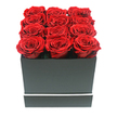 The photograph shows a luxurious cluster of twelve preserved ruby red roses presented in a matte black square gift box, captured in clear, even light that highlights every texture and curve. Each rose is in perfect, full bloom, with velvety petals spiralling inward in tight, layered whorls that create a plush, almost cushion-like effect when seen together. The flowers are arranged in a precise three-by-four grid across the top of the box, their heads nestled shoulder to shoulder so that a continuous field of deep crimson stretches from edge to edge, the blooms sitting just proud of the rim. The rich red tones vary subtly, with darker shadows pooling in the petal folds and slightly brighter highlights catching the outer edges, giving the roses depth and realism. The box beneath them is a minimalist black cube with crisp lines and a smooth, non-reflective finish, refined by a narrow white stripe that runs horizontally around its lower section like a chic detail on a high-end gift from a Stanmore boutique. Set against a pure white backdrop, there are no other elements to divert attention-only the strong graphic contrast of red, black, and white. From the standpoint of a local Stanmore florist accustomed to creating elegant, meaning-filled gifts for homes near Belmont Circle and along Marsh Lane, this composition feels ideal for declarations of love, thoughtful anniversaries, or sophisticated Valentine's Day surprises. The preserved nature of the roses suggests enduring sentiment, a lasting reminder of a special moment delivered directly to a loved one's door.